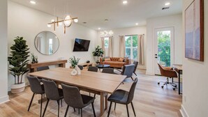 A modern living room with a large wooden dining table, comfortable seating, a flat-screen TV, and a large window with curtains.