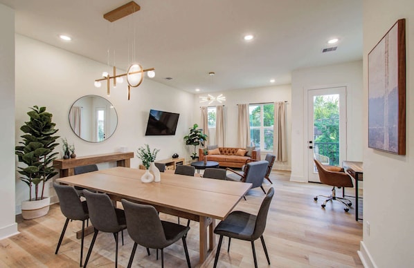 A modern living room with a large wooden dining table, comfortable seating, a flat-screen TV, and a large window with curtains.