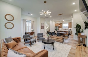 A modern living room with a sofa, chairs, a coffee table, and a dining area with a table and chairs. There is a kitchen in the background with cabinets and a refrigerator.