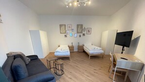 A modern, well-lit room with a wooden floor, two single beds, a sofa, a desk with a chair, and a TV.
