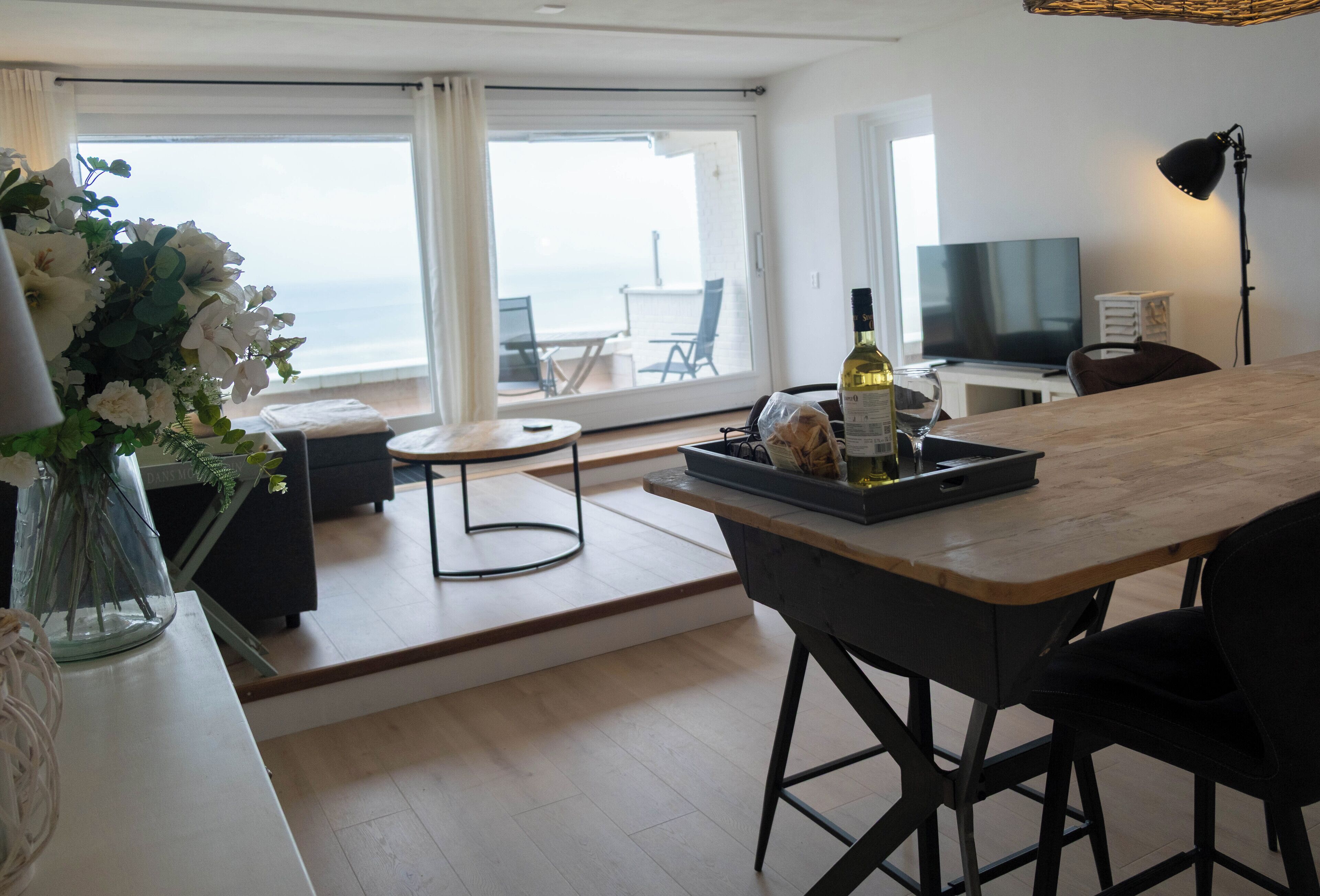 Quarto de hotel moderno com uma janela grande que oferece vista para o mar, área de refeições com um banco de bar, TV de tela plana e uma luminária.