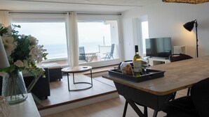 A modern hotel room with a large window offering a view of the sea, a dining area with a bar stool, a flat-screen TV, and a lamp.