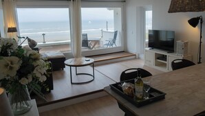 A modern living room with a large window overlooking the beach, a flat-screen TV, and a dining area with a tray of food and drink.
