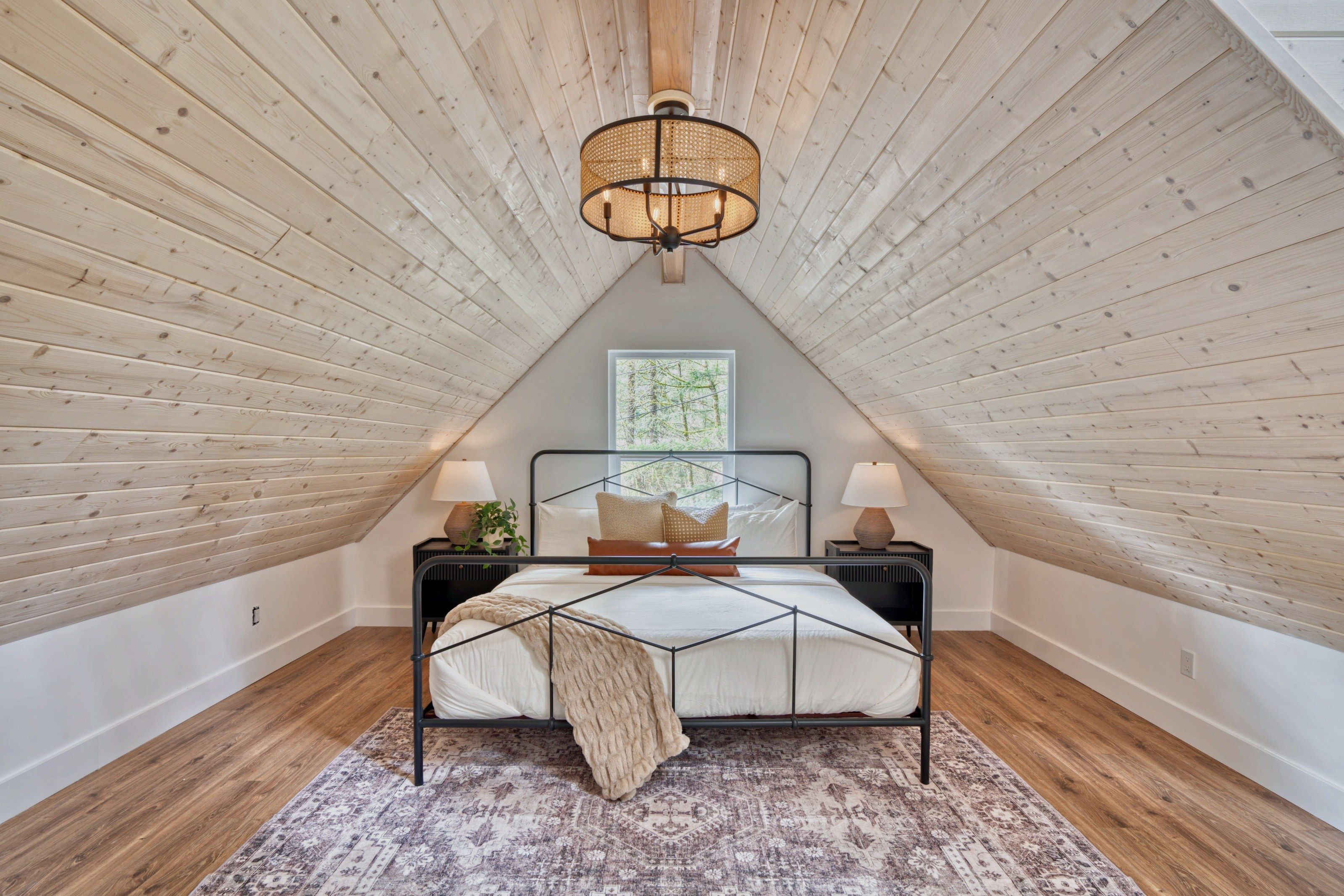 A bedroom with a sloped ceiling, a bed with a geometric headboard, two bedside tables with lamps, a patterned rug, and a window with a view of trees.