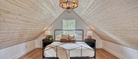 A bedroom with a sloped ceiling, a bed with a geometric headboard, two bedside tables with lamps, a patterned rug, and a window with a view of trees.