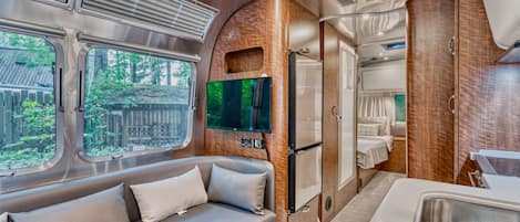 A compact RV interior with a sofa, a flat-screen TV, a refrigerator, and a small kitchenette.