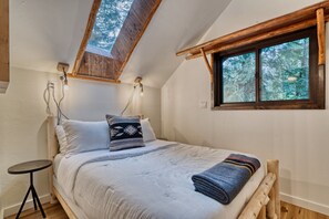 A bedroom with a sloped ceiling, a skylight, a bed with pillows, a bedside table, and a window with a view of trees. - Log Cabin Compound | 6BR/5.5BA | 2 Hot Tubs (Mount Hood Village)