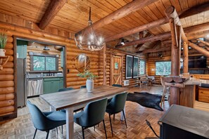 A log cabin interior with a dining area, kitchen, and a large window overlooking a forest. - Winter Log Cabin Compound • 2 Hot Tubs • Sleeps 14 (Mount Hood Village)