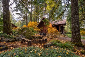 A rustic cabin surrounded by a forest with a wooden table and benches in the foreground. - Log Cabin Compound | 6BR/5.5BA | 2 Hot Tubs (Mount Hood Village)