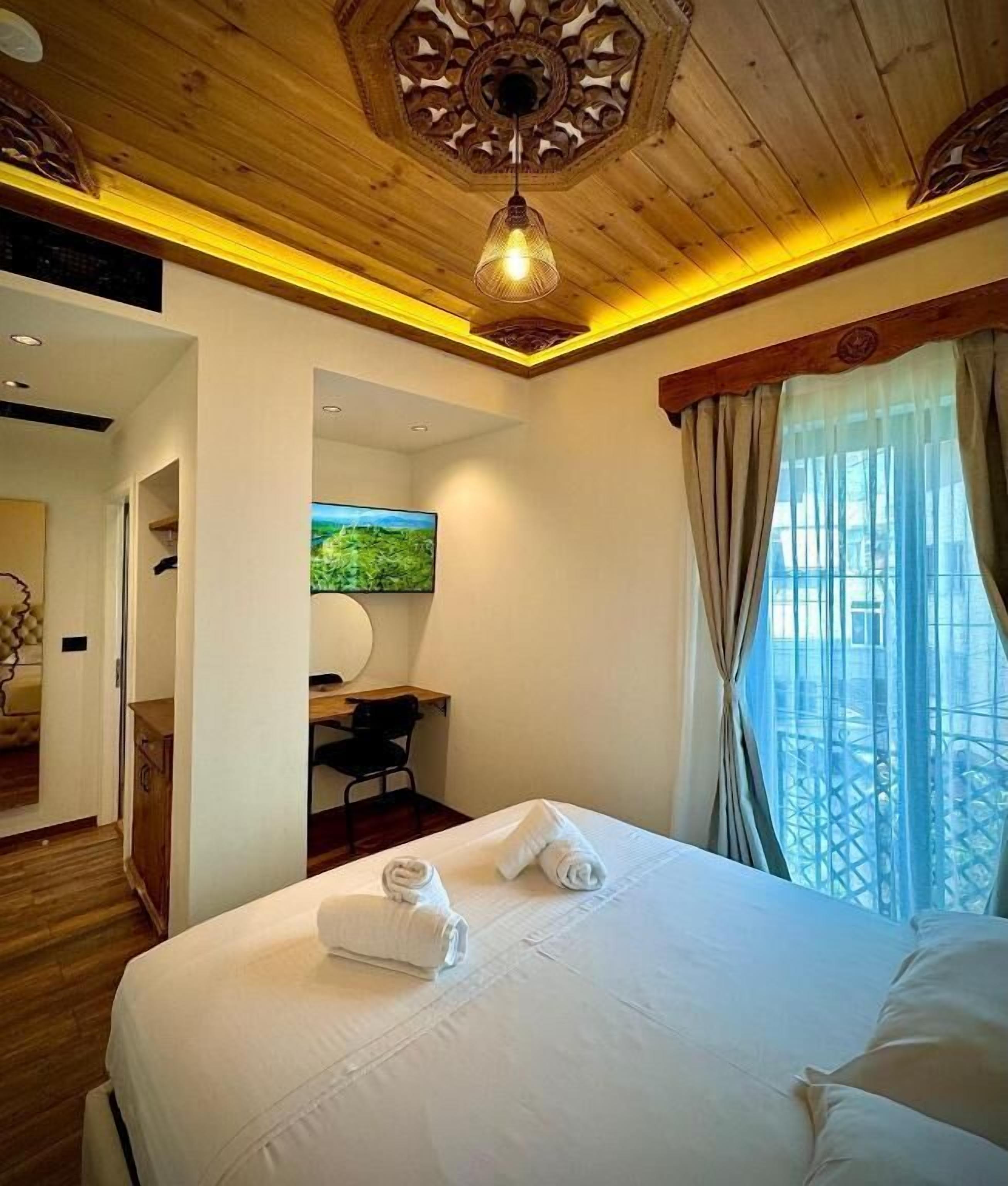 Camera d'albergo con soffitto in legno, un letto con lenzuola bianche, una scrivania e un'ampia finestra con tende.