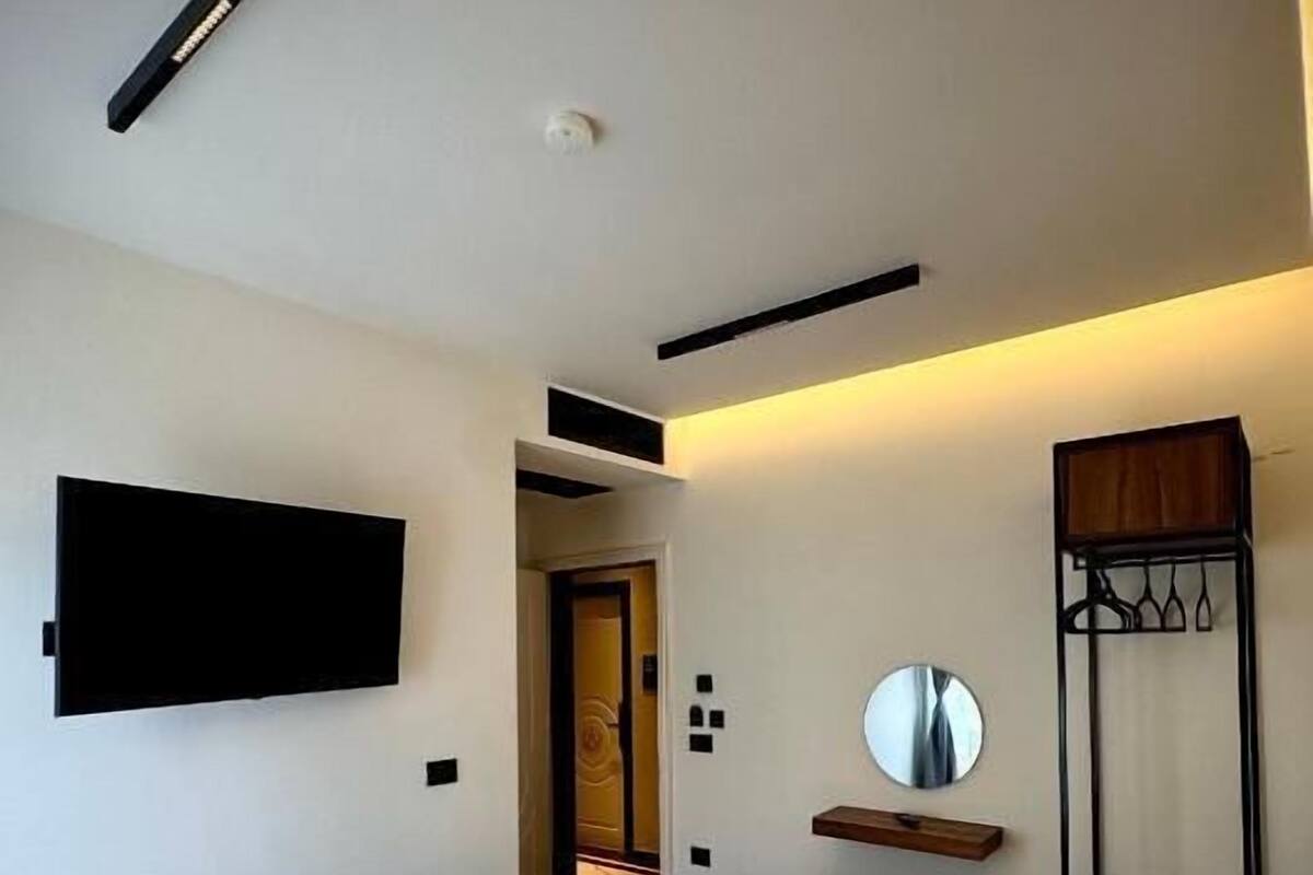 A modern hotel room with two single beds, a flat-screen TV mounted on the wall, a bedside table with a mirror, and a coat rack.