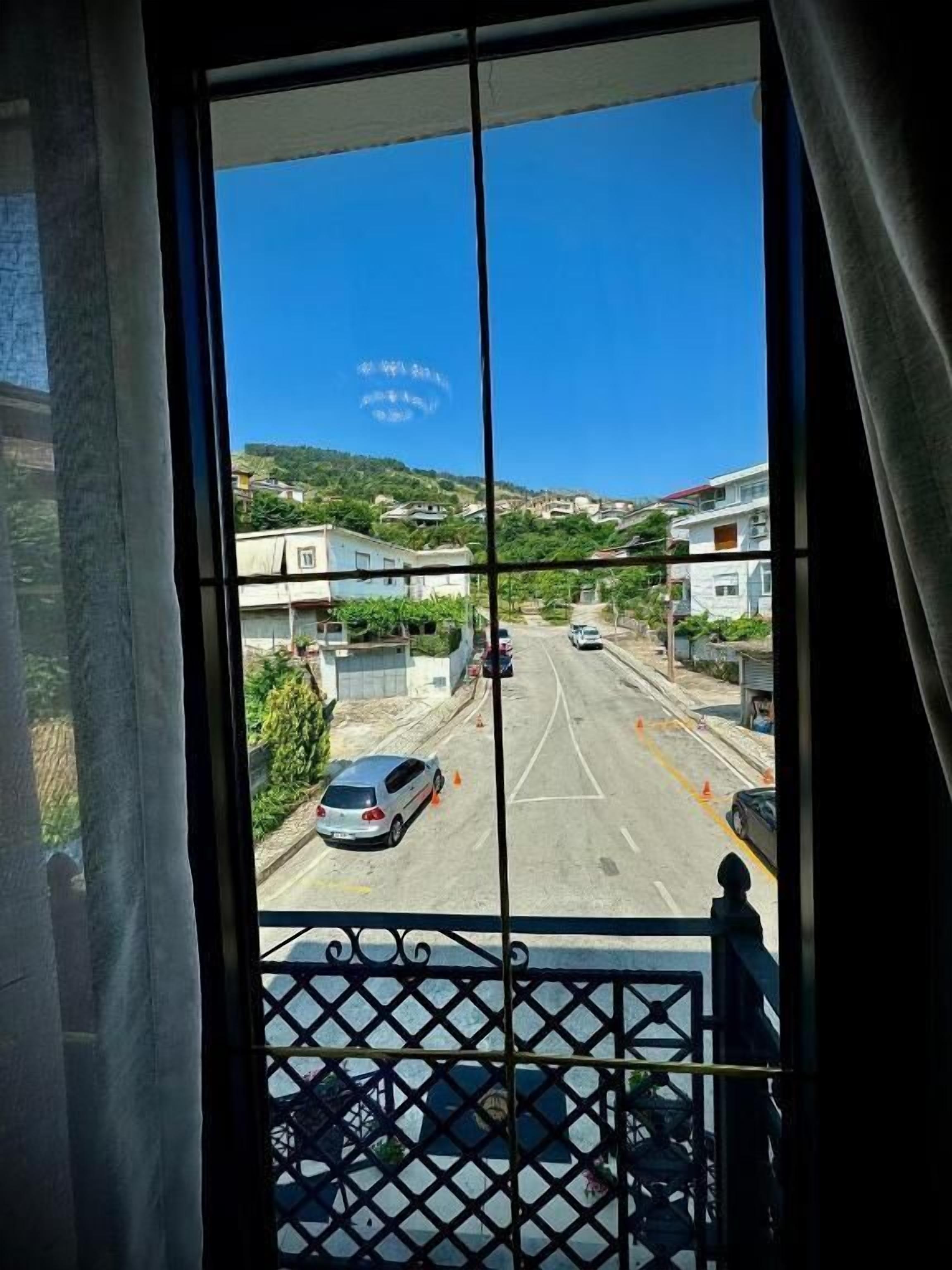 Vista desde una ventana con celosía metálica, que da a una calle con coches y edificios, bajo un cielo azul despejado.