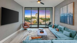 A spacious living room with a large TV, a ceiling fan, a sofa set, a coffee table, and a view of a cityscape through large windows.