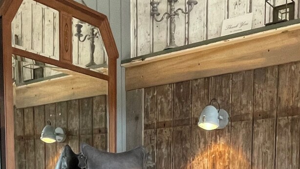 Ein gemütliches Zimmer mit einer Holzwand, einem Spiegel, einem schwarzen Stuhl mit flauschigem weißen Kissen und einem kleinen Tisch mit Lampe.