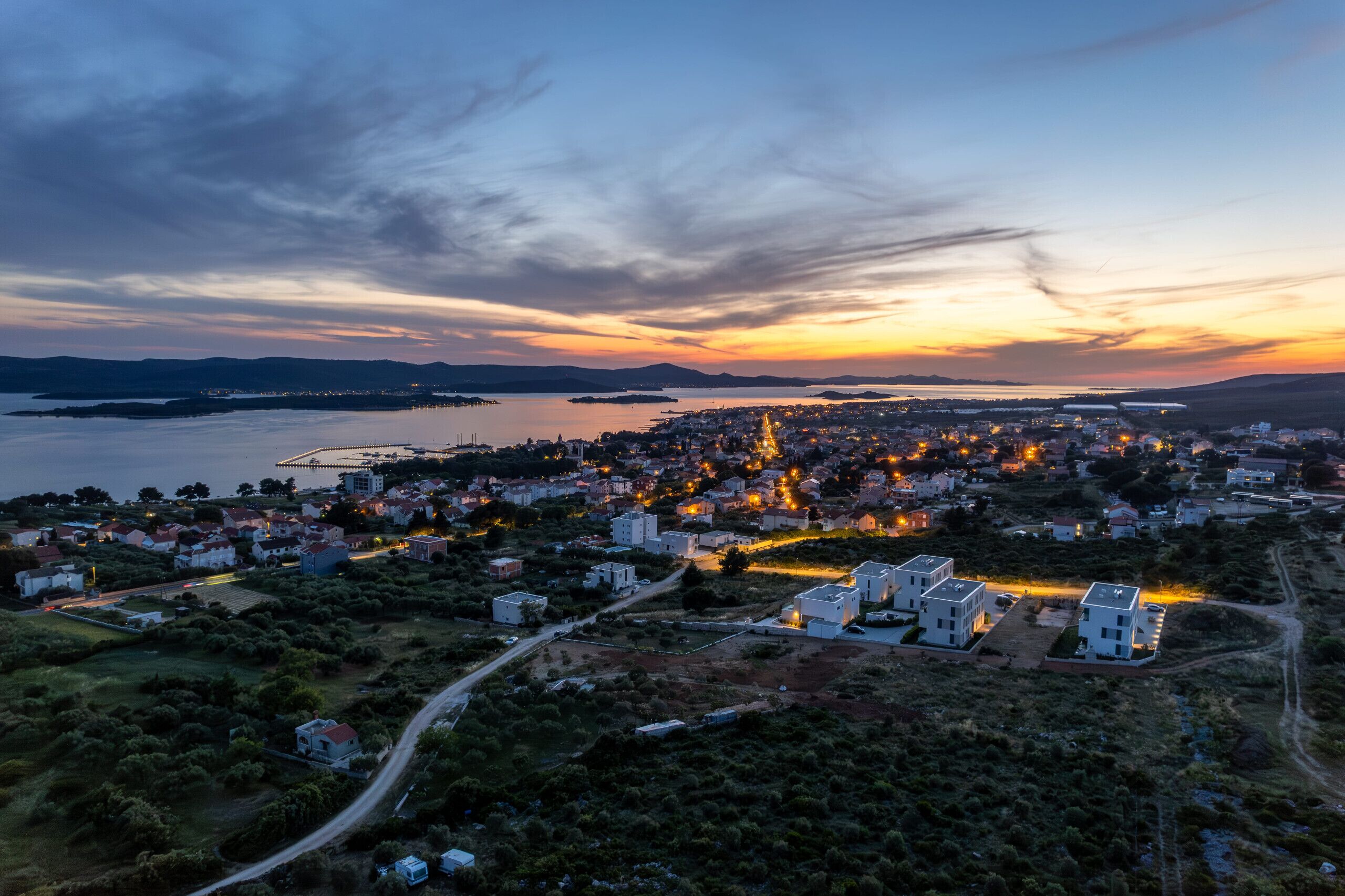 An aerial view of a coastal town at dusk with buildings, roads, and a body of water.