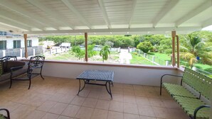 Un balcon abrité avec un sol carrelé, deux chaises en métal, une table en métal et un banc vert.