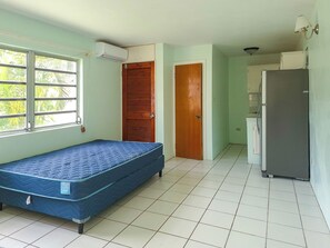 Ein kleiner, kompakter Raum mit einem Einzelbett, einer Holztür, einem Kühlschrank und einem Fenster mit Blick auf Palmen.