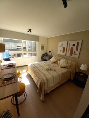 Un dormitorio con cama, escritorio y silla. Hay una ventana que proporciona luz natural y la habitación tiene una decoración en tonos neutros.