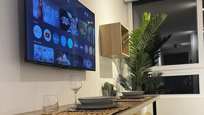 Un comedor moderno con un televisor de pantalla plana montado en la pared que muestra diversos servicios de transmisión. Hay una mesa de madera con un mantel individual, un control remoto y un vaso. Dos sillas amarillas están ubicadas a la mesa, y se ve una planta en maceta al fondo.