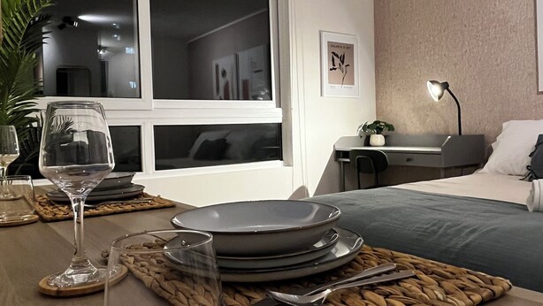 Habitación de hotel moderna con comedor que incluye una mesa de madera dispuesta con platos, vasos y cubiertos. Un ventanal ofrece vistas al cielo nocturno.