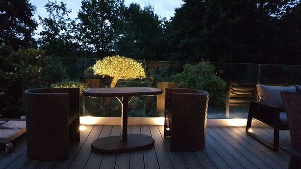 Terraza exterior con una mesa redonda y dos sillas, rodeada por un suelo de madera y una barandilla de cristal.