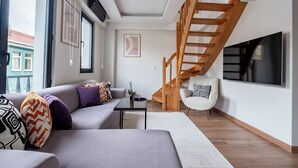 Un salon moderne avec un canapé gris, un fauteuil blanc, un escalier en bois et un grand téléviseur fixé au mur.