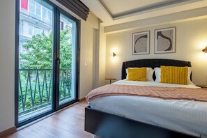 A bedroom with a bed, a nightstand, a window with a view of greenery, and two wall-mounted lights.