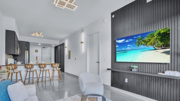Ett modernt vardagsrum med en stor platt-TV, en soffa, ett soffbord och en matplats med stolar. Köket syns i bakgrunden.