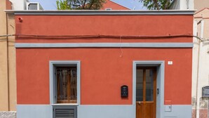 Ein Gebäude mit roter Fassade, zwei Fenstern mit Metallgitter und einer Holztür sowie einem kleinen Briefkasten.