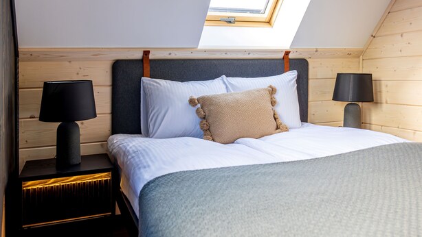 A bedroom with a sloped ceiling, a skylight, a bed with white bedding, two bedside tables with lamps, and a wooden headboard. - RentPlanet - Wierzchołki (Czarny Dunajec)