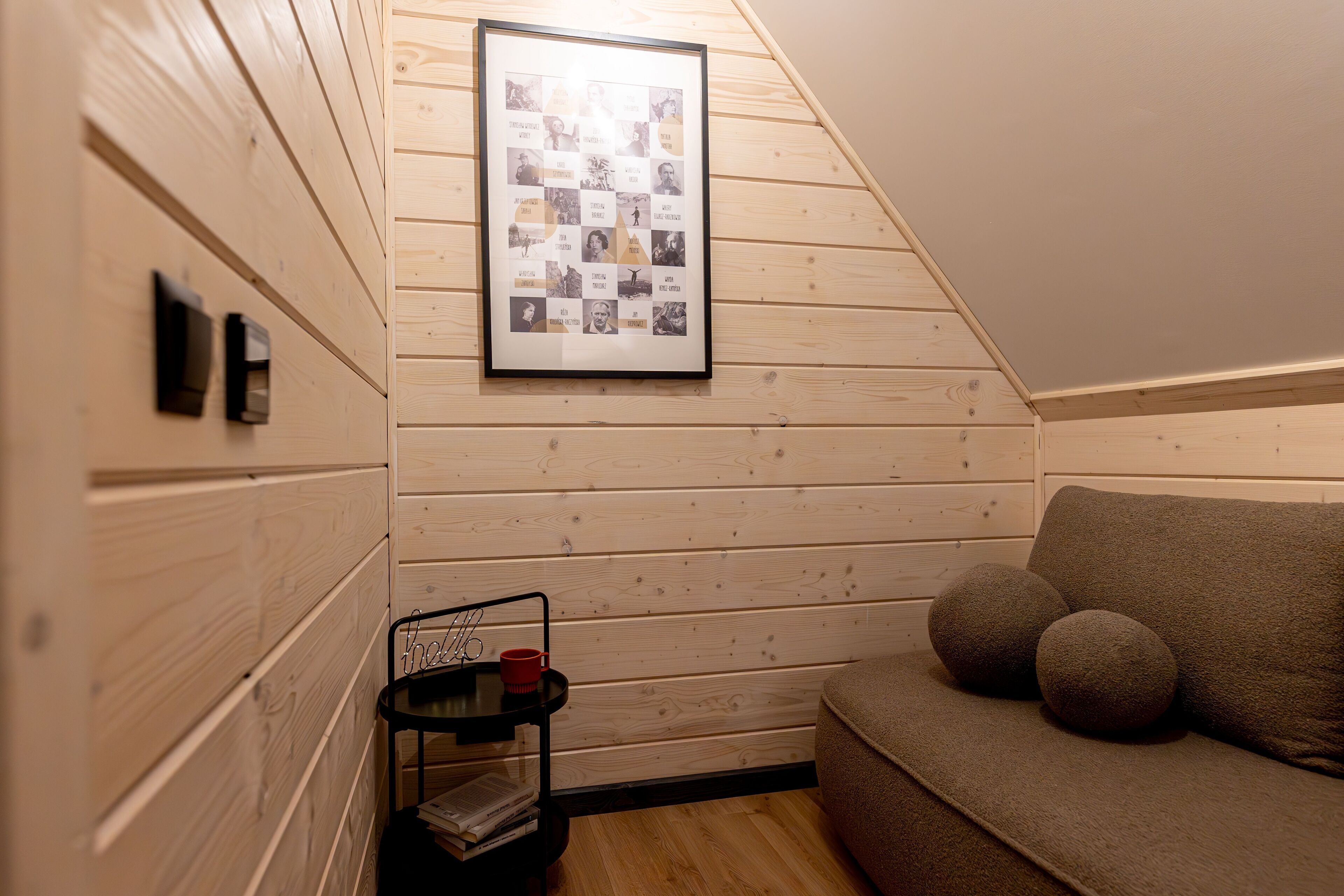 Een knus vertrek met schuine wanden, houten paneelbekleding, een lijstwerkcollages aan de muur, een klein zwart bijzettafeltje met een rode beker en een bruine bank met ronde kussens.