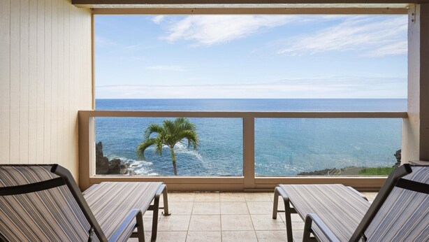 A balcony with two striped chairs, a view of the ocean, and a framed picture of a palm tree and rocky shore.