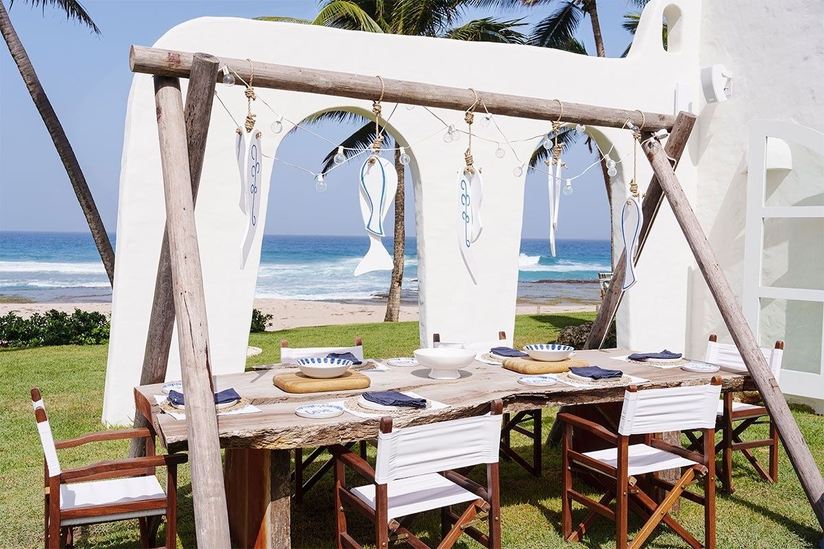 An outdoor dining area with a view of the ocean, featuring a rustic wooden table and chairs, white walls, and hanging decorations.
