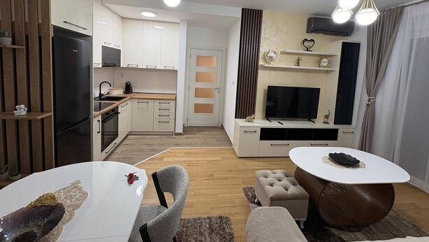 A modern kitchen with white cabinets, a black refrigerator, and a dining area with a white table and grey chairs. - I&I Apartments 36 (Kragujevac)