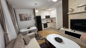A modern living room with a sofa, coffee table, and TV. There is a kitchen area with cabinets and a refrigerator. - I&I Apartments 36 (Kragujevac)