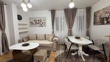 A modern living room with a beige sofa, a round coffee table, and a dining table with chairs. There are curtains, a painting, and a clock on the wall.