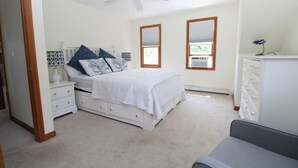 A bedroom with a bed, nightstands, a dresser, a ceiling fan, and two windows with blinds. - Sunny Cape Cod Poolside Haven (2407) (Truro)