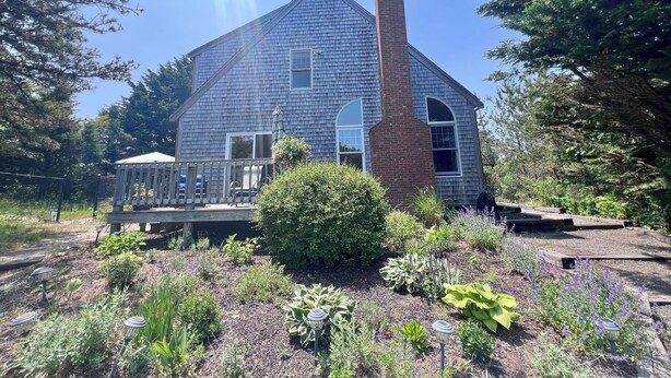 A two-story house with a large chimney, a covered porch, and a garden with various plants. - Sunny Cape Cod Poolside Haven (2407) (Truro)