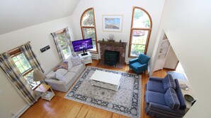 A spacious living room with a fireplace, two sofas, a television, and large windows. - Sunny Cape Cod Poolside Haven (2407) (Truro)