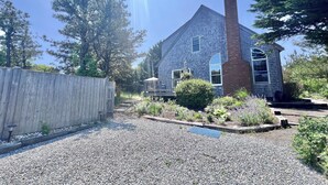 A house with a gravel driveway, a wooden fence, and a garden with various plants. - Sunny Cape Cod Poolside Haven (2407) (Truro)