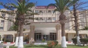A hotel with a central entrance, palm trees, and a sign in a foreign language. - Hôtel Club Les Colombes (Hammamet)