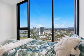 Une chambre avec une grande fenêtre, un lit recouvert d’une couvre-lit à motifs floraux et une vue sur le paysage urbain.