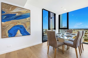 A modern dining area with a large abstract painting, a glass table, and chairs, with a city view through large windows.