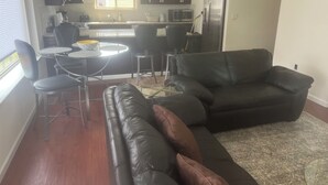 A living room with a black leather sofa, a dining area with a small round table and chairs, and a kitchen with dark cabinets and stainless steel appliances.