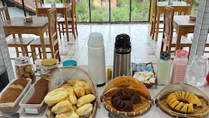A buffet setup with various food items and utensils, surrounded by wooden chairs and tables, with a view of greenery outside. - Hotel Lumiar (Nova Friburgo)