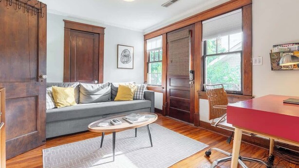 A room with a grey sofa, a wooden door, a red desk, and a window with blinds. - Boscobel Bungalow - Walkable to Shelby Park (Nashville)
