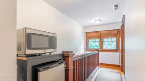 A narrow hallway with a microwave on a black cabinet, a wooden railing, and a window with blinds. - Boscobel Bungalow - Walkable to Shelby Park (Nashville)