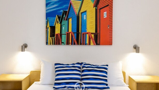 Een hotelkamer met een bed, twee nachtkastjes en een kleurrijk schilderij van strandhuisjes.