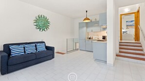 Een moderne woonkamer met een bank en decoratief wandkunstwerk, met doorgang naar een keuken met een eettafel en stoelen.