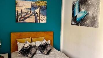 Une chambre avec un mur de couleur turquoise, une tête de lit en bois et un lit recouvert d’une literie à motifs. Deux tableaux ornent le mur : l’un représente une scène de plage et l’autre, un papillon bleu.